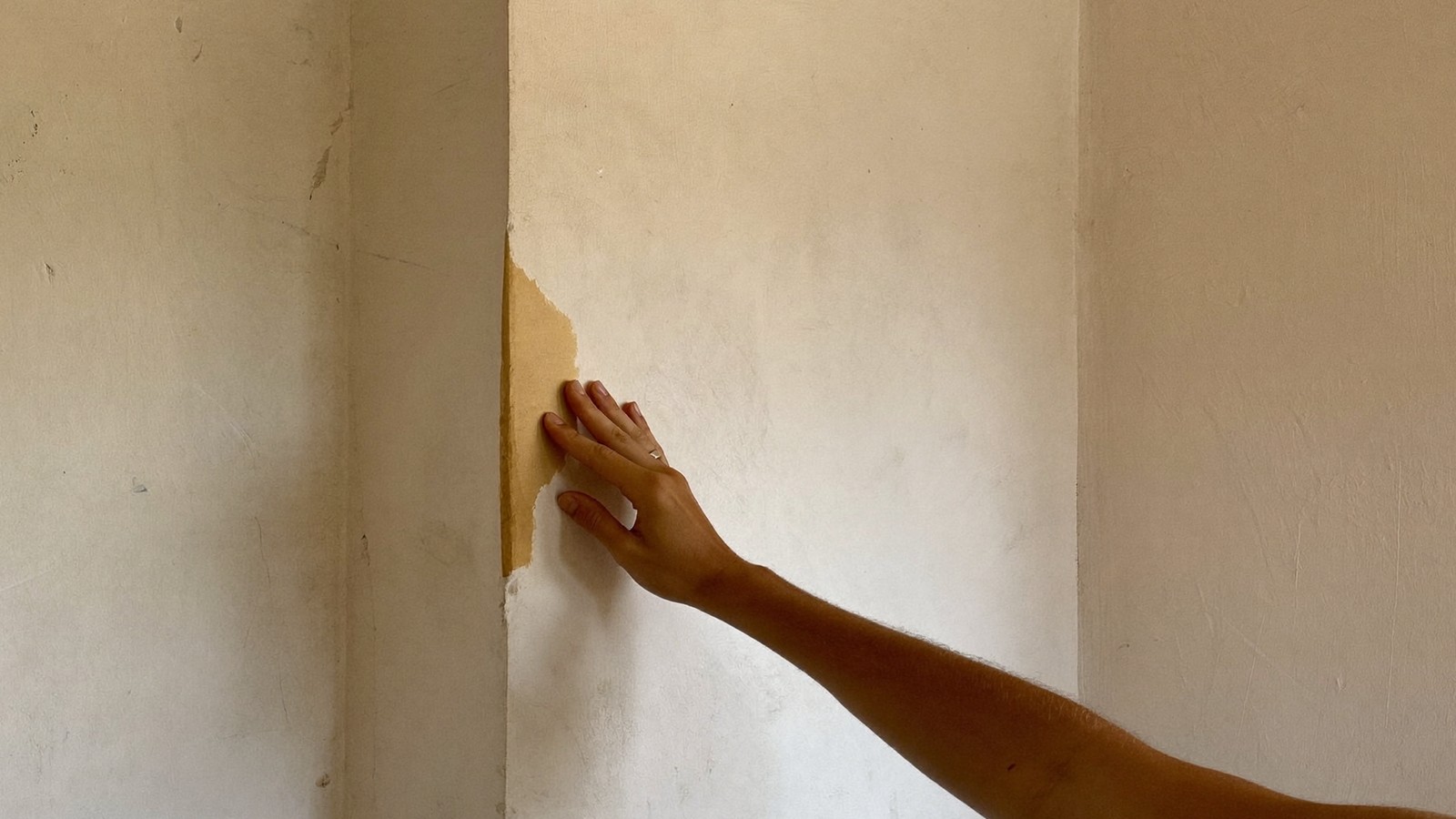 Mano tocando el borde de una zona dañada por humedad en una pared de yeso clara